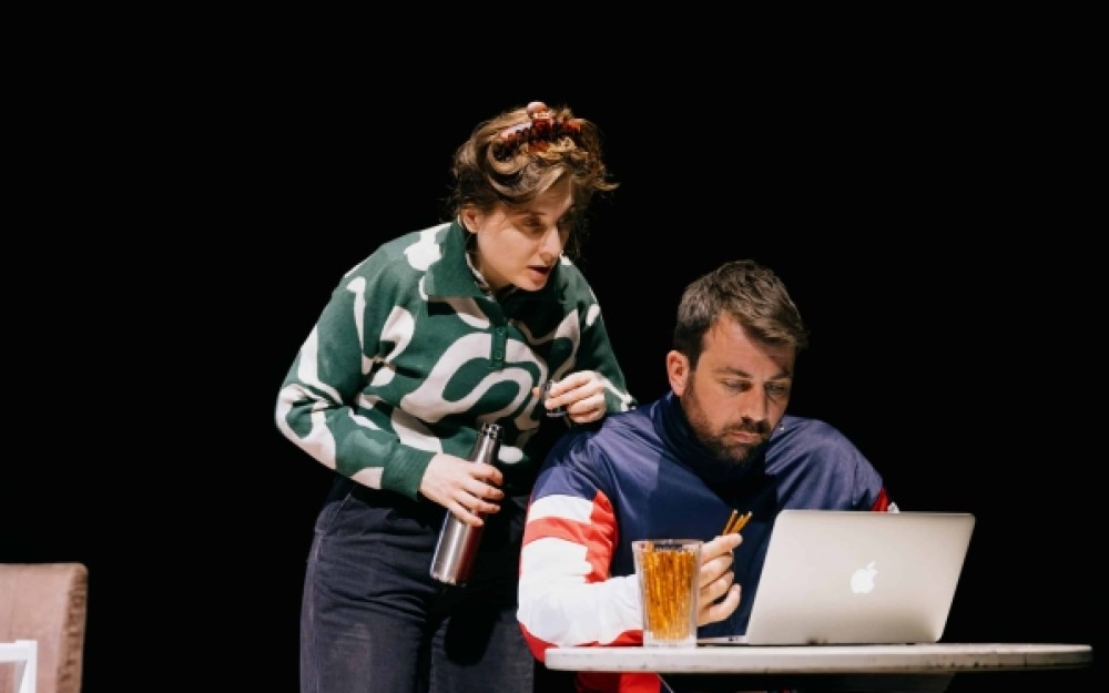 Das Bild zeigt eine Szenenausschnitt aus dem Stück "Die Quelle". Zu sehen ist ein Mann,
                                                der vor seinem Laptop an einem Tisch sitzt. Links von ihm steht eine Frau