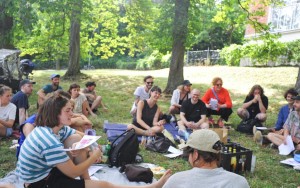 Eine Gruppe Menschen, Mitglieder des Kollektiv Kaorle, sitzt
                                 vor dem Pavillon 21 in der Wiese und bespricht wie es weiter geht.