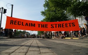 Das Bild zeigt eine
                                 gepflasterte Straße, über die ein roter Banner mit dem Schriftzug "Reclaim the streets" gespannt ist.