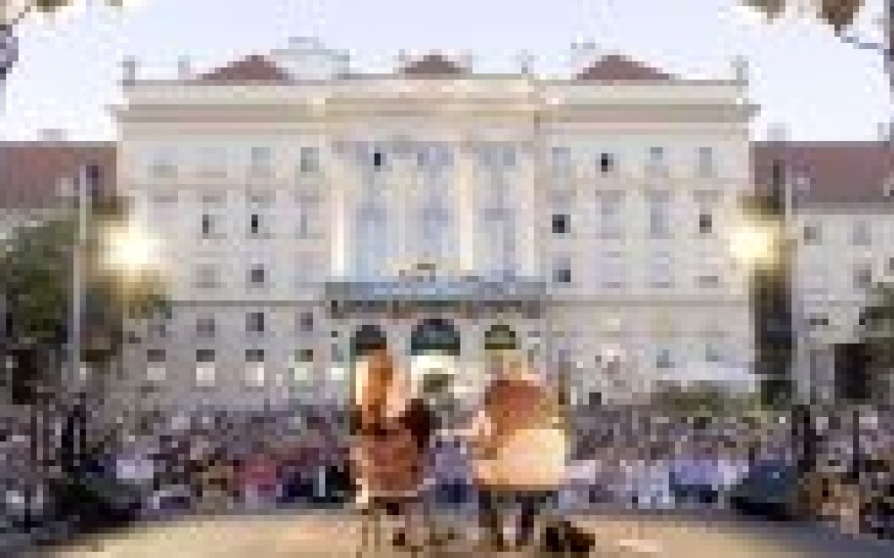 Im Vordergrund die Bühne von hinten, auf der Bühne sitzen
                                                2 Personen, im Hintergrund die Fassade des Hauptgebäudes vom Museumsquartiers, dazwischen zahlreiche Menschen, die der Veranstaltung
                                                beiwohnen.